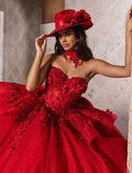 A woman models a dramatic deep red ball gown with a sweetheart neckline, 3D floral appliqués, and a matching red hat with large floral accents. She is posed against a beige stone wall, showcasing the dress's full voluminous skirt.
